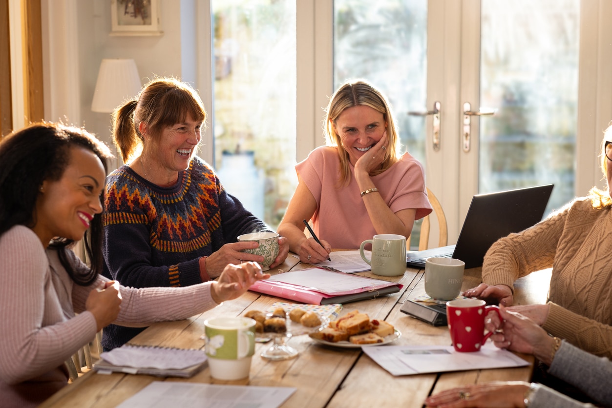 A medium shot of a group of women having a community meeting together. They are all sitting down around a table with treats and hot drinks. They are taking notes while discussing and laughing.